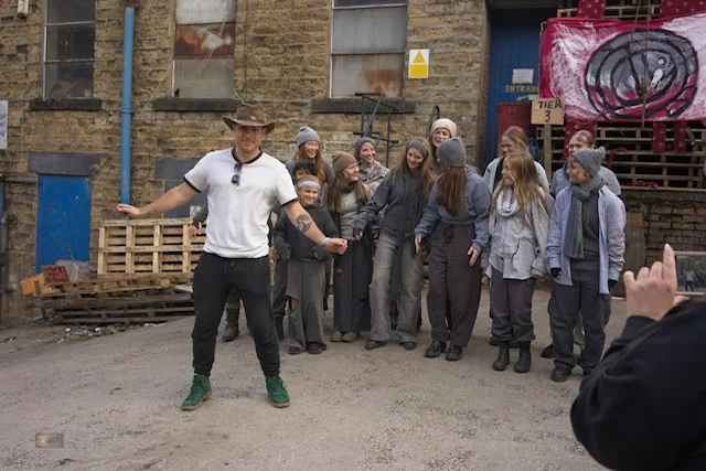 A man in a wide-brimmed hat and white T-shirt stands in front of a group of young cast members in grey costumes in a yard between old stone buildings, while someone films on a phone