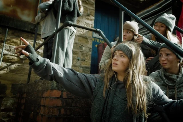 A girl in a grey beanie and scarf points into the distance on a stone stairway, with other young cast members in grey costumes behind her